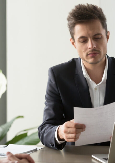 Company leader reading contract, attentively studying text of agreement, doubting some deal conditions while sitting at desk with business partner. Pensive CEO examining financial document or plan